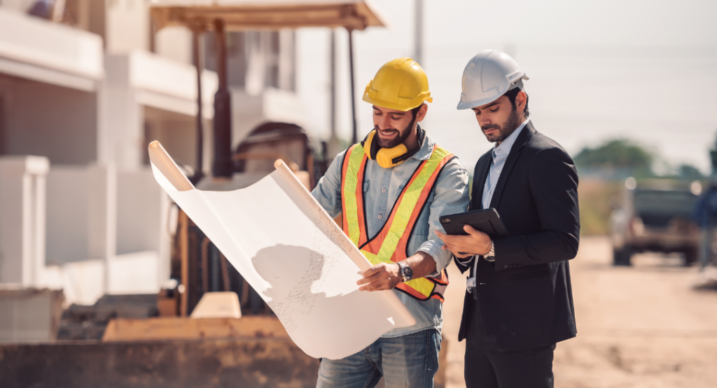 Um engenheiro e um gerente de obras em capacetes, analisando plantas e um tablet num canteiro de obras, focando no gerenciamento de obras eficiente.
