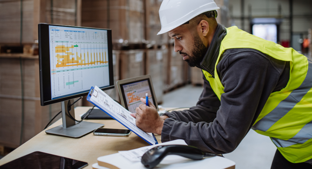 Profissional de colete e capacete revisando dados e planilhas em um monitor, utilizando um sistema de segurança do trabalho para monitoramento e controle em um ambiente industrial.