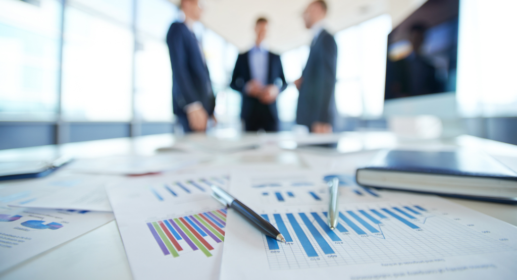 Documentos de relatórios de não conformidades com gráficos sobre a mesa e executivos ao fundo.